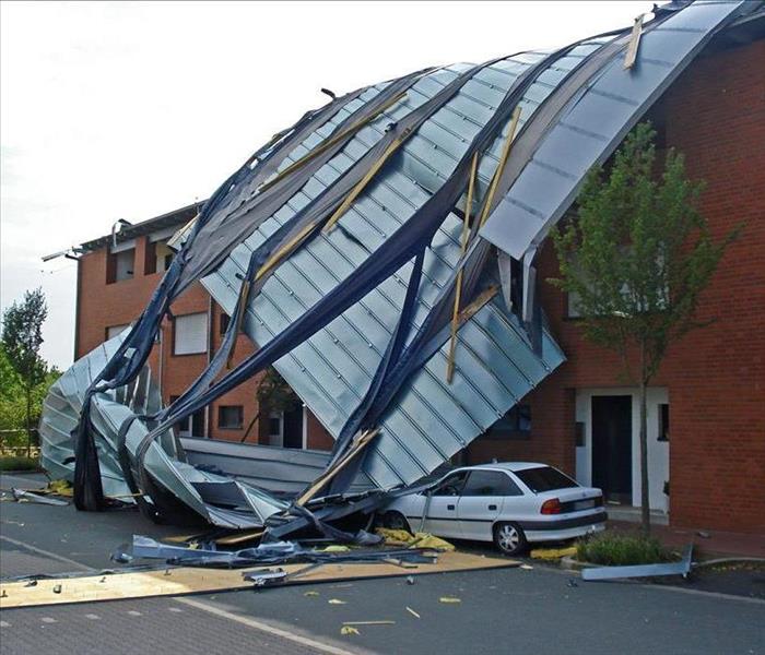 Roof damaged by wind that has fallen on a white car outside a building.