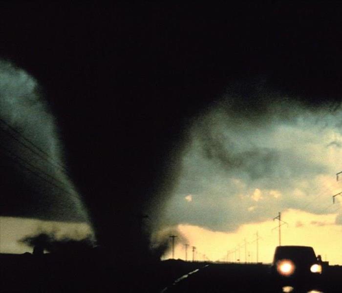 Black tornado funnel cloud on a road next to a car with telephone poles in the background.